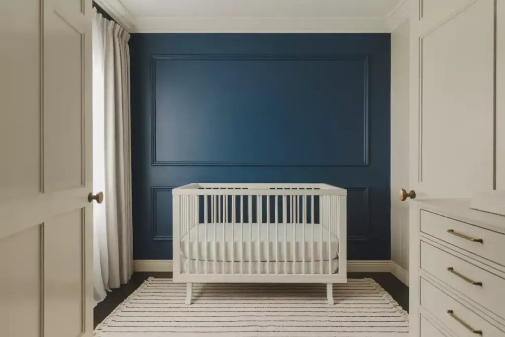 White crib on a striped rug against a dark blue accent wall in a modern, minimal nursery with white curtains.