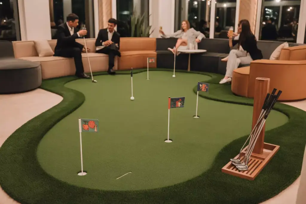 Four people relax around an indoor mini golf green with flags and golf clubs, chatting and drinking.