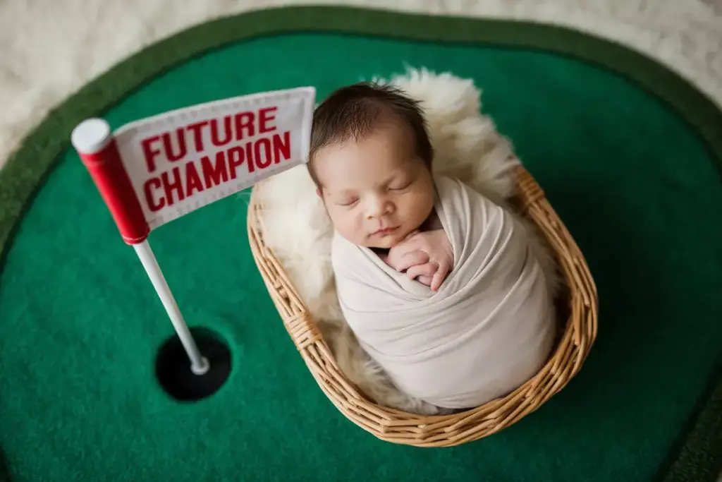 Baby wrapped in a blanket sleeps in a basket next to a golf flag that says “Future Champion.”.
