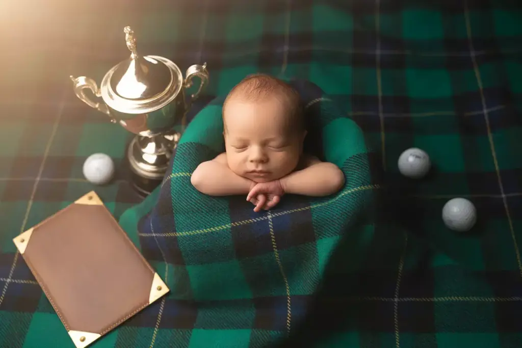 Newborn baby wrapped in a green plaid blanket beside a trophy, golf balls, and a blank plaque.
