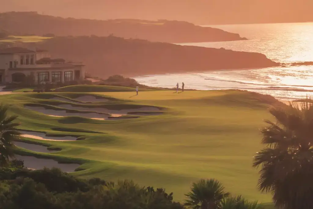 Golf course by the ocean at sunset with silhouettes of two people and a clubhouse in the distance.
