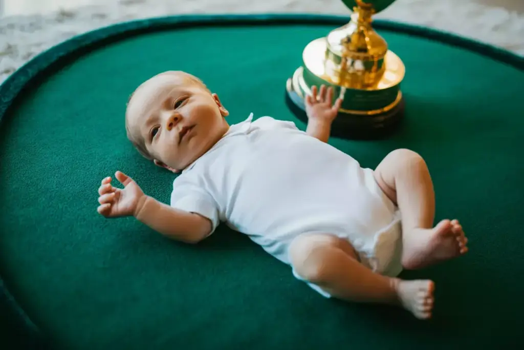 Baby in a white onesie lying on a green mat next to a large gold trophy.
