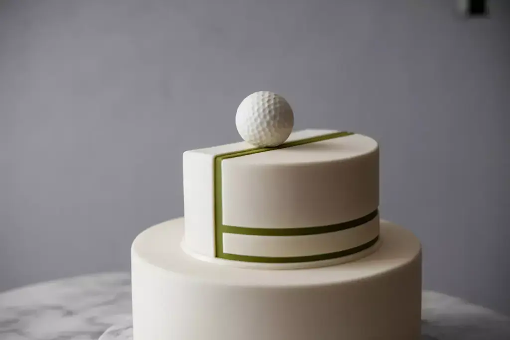 Smiling golf ball cartoon figure with a club stands on a golf-themed cake decorated with flags and holes.