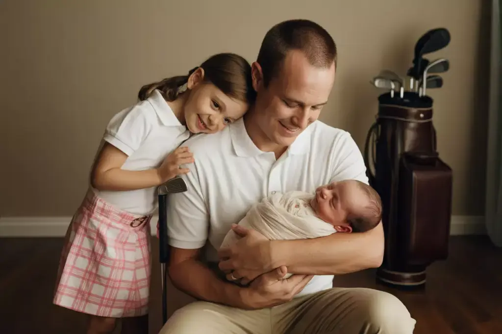 A father holds a swaddled newborn while a young girl hugs him, with golf clubs in the background.