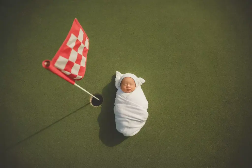 A swaddled baby lies on a golf green beside a hole with a red checkered flag.