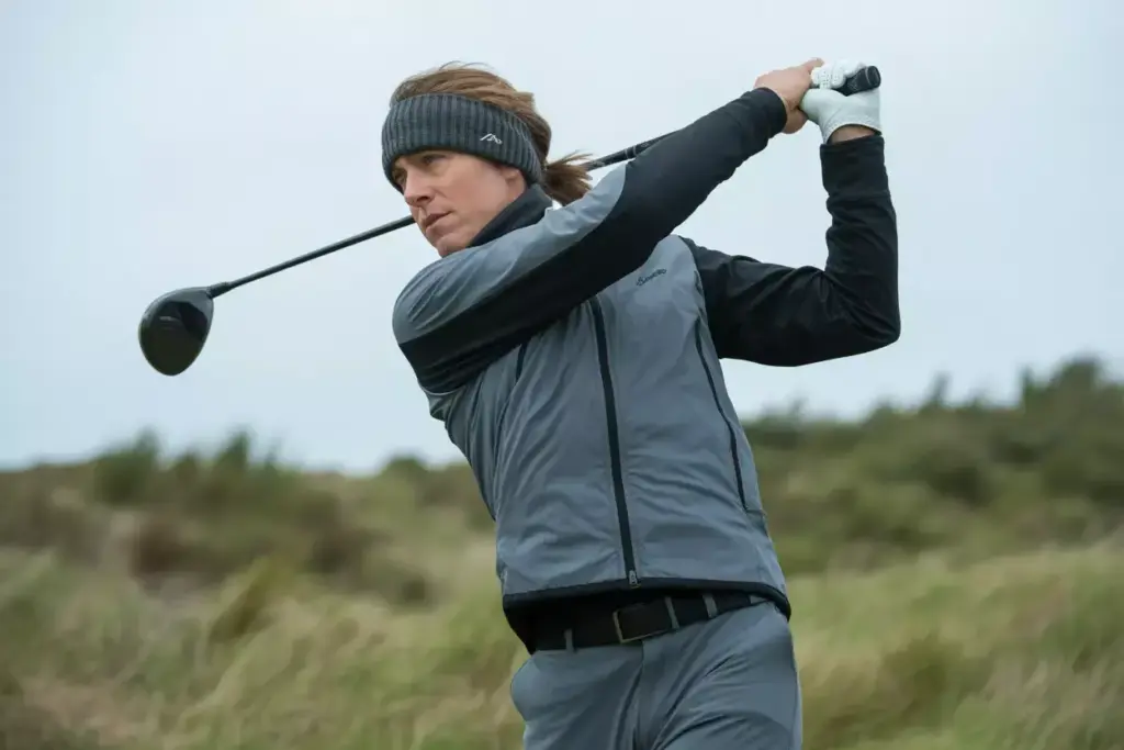 A Golfer Mid Swing In A Wind Resistant Soft Shell Jacket And Thermal Leggings On A Breezy Course