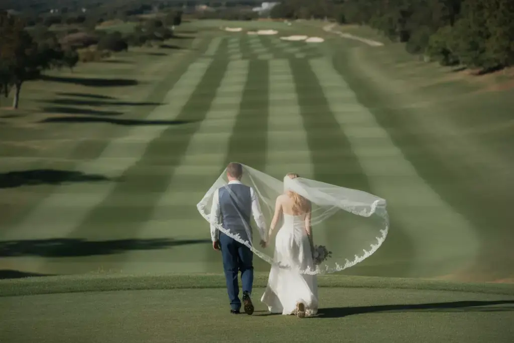 Couple Walking Hand In Hand Down Expansive Manicured Fairway With Brides Veil Flowing In Breeze